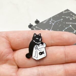 A black cat enamel pin sitting inside a white grocery bag that has a small fish icon on the front.