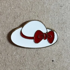 A white wide-brimmed sun hat enamel pin with a vibrant red ribbon and bow, finished with gold edging.