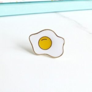 A simple, minimalist enamel pin of a sunny-side-up fried egg with a bright yellow yolk and an irregular white border.