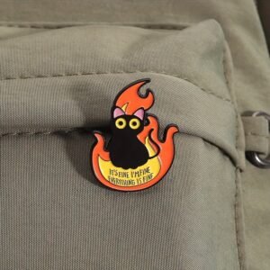 Hard enamel pin of a black cat with big yellow eyes sitting in orange flames with the text "IT'S FINE I'M FINE EVERYTHING IS FINE".