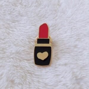 A gold-plated enamel pin shaped like a black lipstick tube with a red tip and a gold heart on the base.
