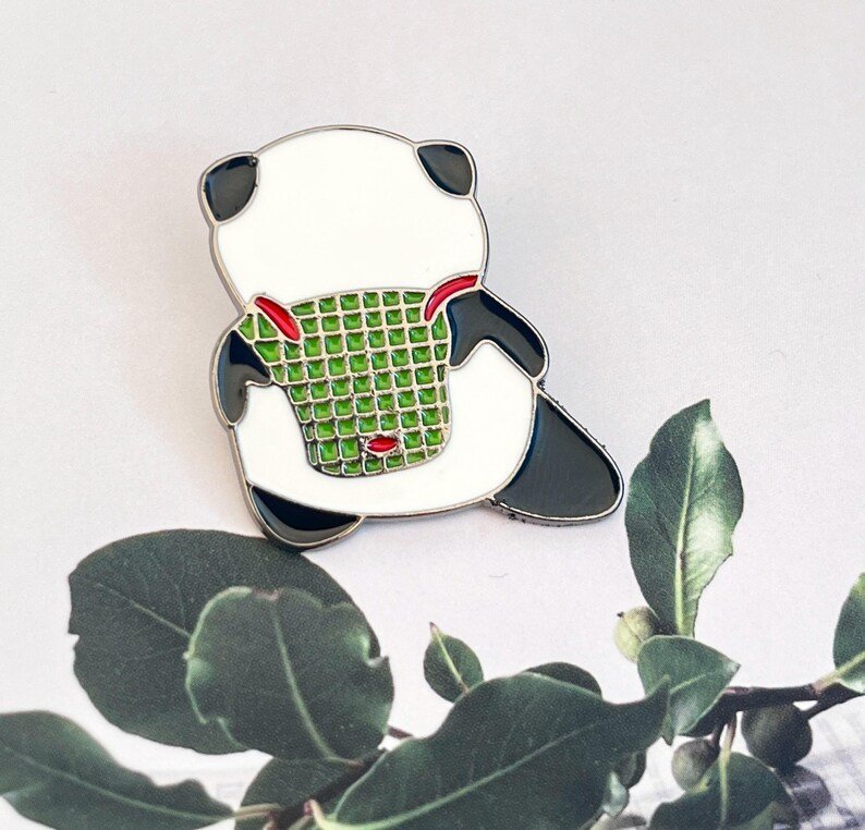 A back-view enamel pin of a panda bear carrying a green woven basket or backpack with red straps.