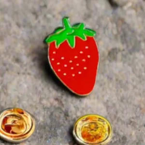 Bright red strawberry enamel pin with a green leafy stem and yellow seed details.