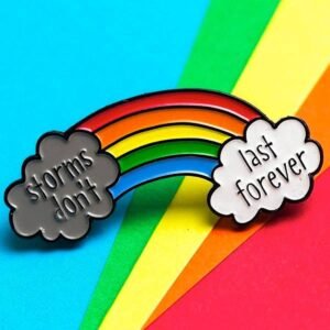 A colorful arc rainbow enamel pin connecting a dark grey rain cloud on the left with a fluffy white cloud on the right, featuring the text "storms don't last forever."