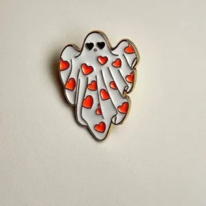 A cute white ghost enamel pin with gold metal edging, covered in a pattern of small red hearts and wearing heart-shaped sunglasses.