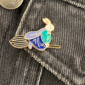 A whimsical enamel pin of a white rabbit wearing a blue and teal wizard outfit riding a broomstick.