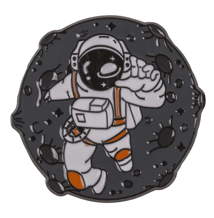 Astronaut-rum-badge – kosmonaut i hvid rumdragt med orange detaljer, der svæver blandt mørke asteroider; emalje-pin med gunmetal-base